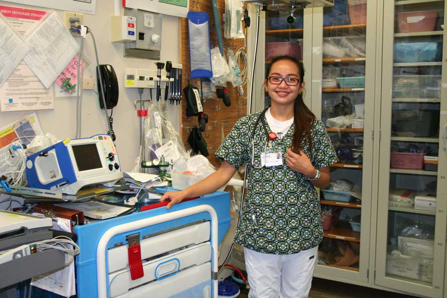 A female nurse in green and white scrubs stands in a room of medical equipment