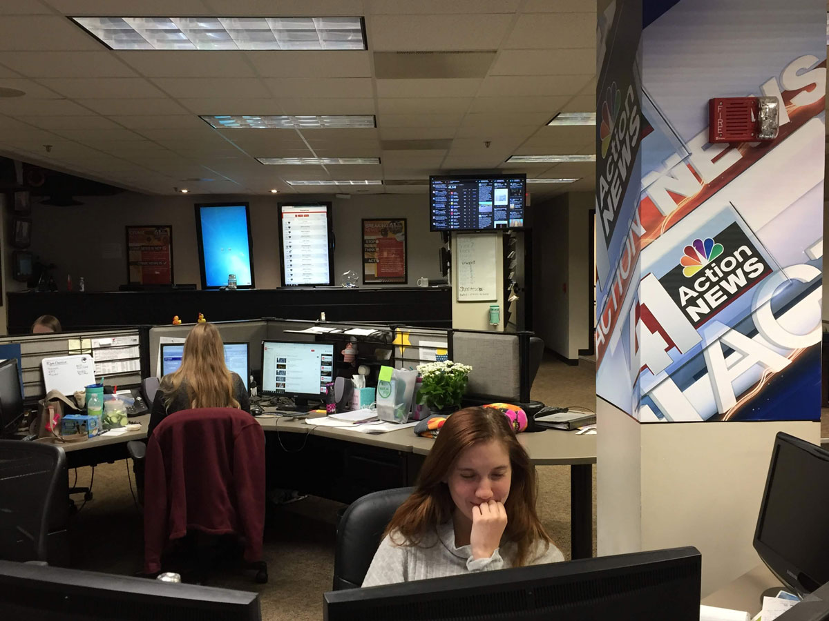 A person with long red hair smiles at a monitor in a TV news room