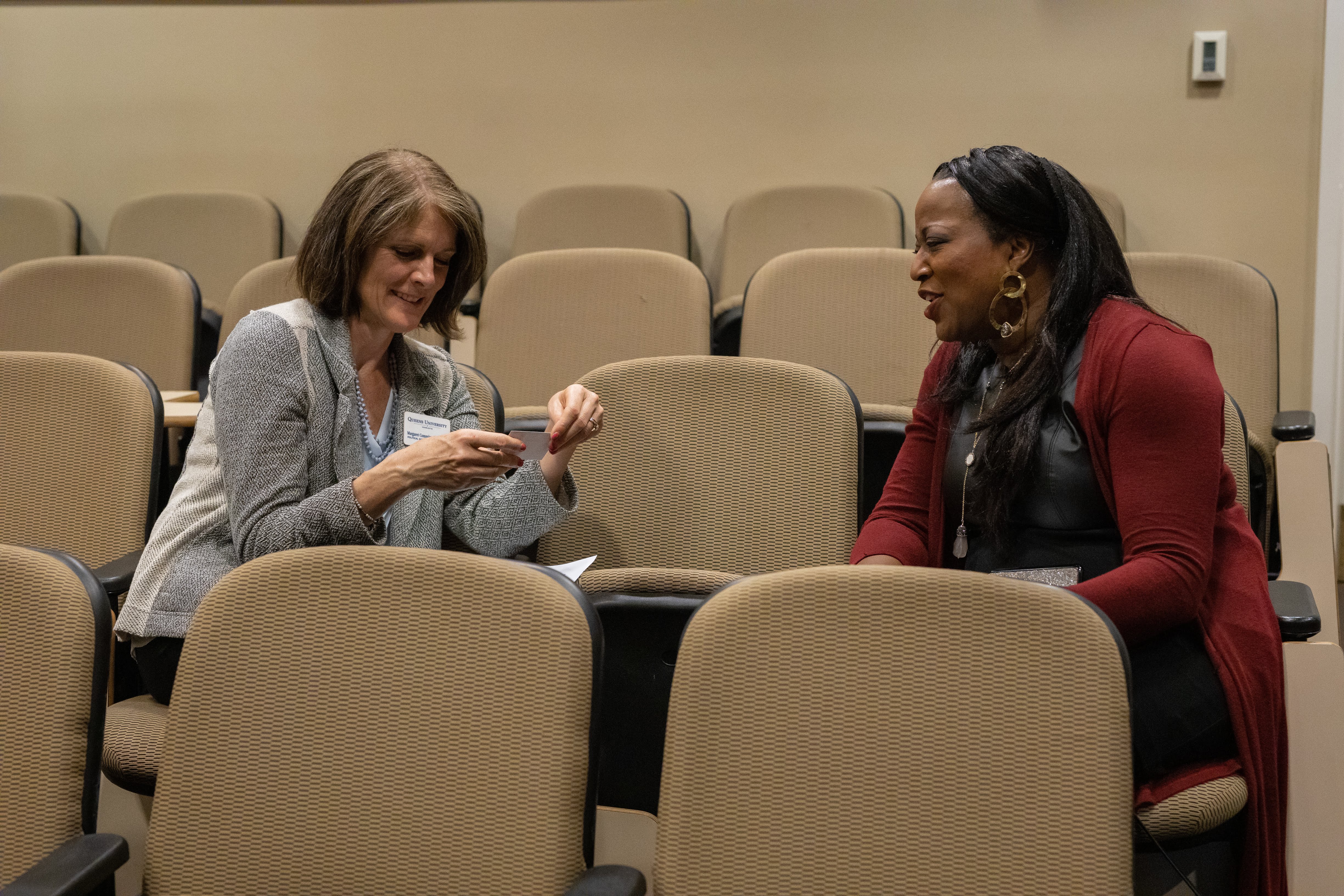 2 people sitting in auditorium beige seating, one seat apart, talking to each other