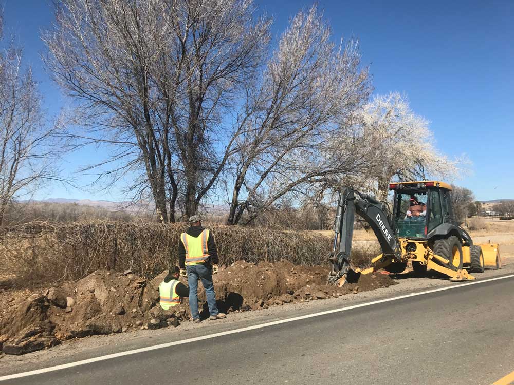 Two workers in yellow reflective vests work in a ditch as another worker operates a hydraulic mining shovel