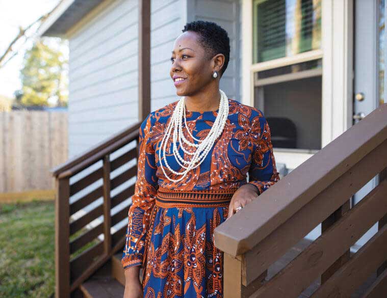 A Black woman wearing a blue and orange dress and a string of pearl necklaces stands outside on a staircase