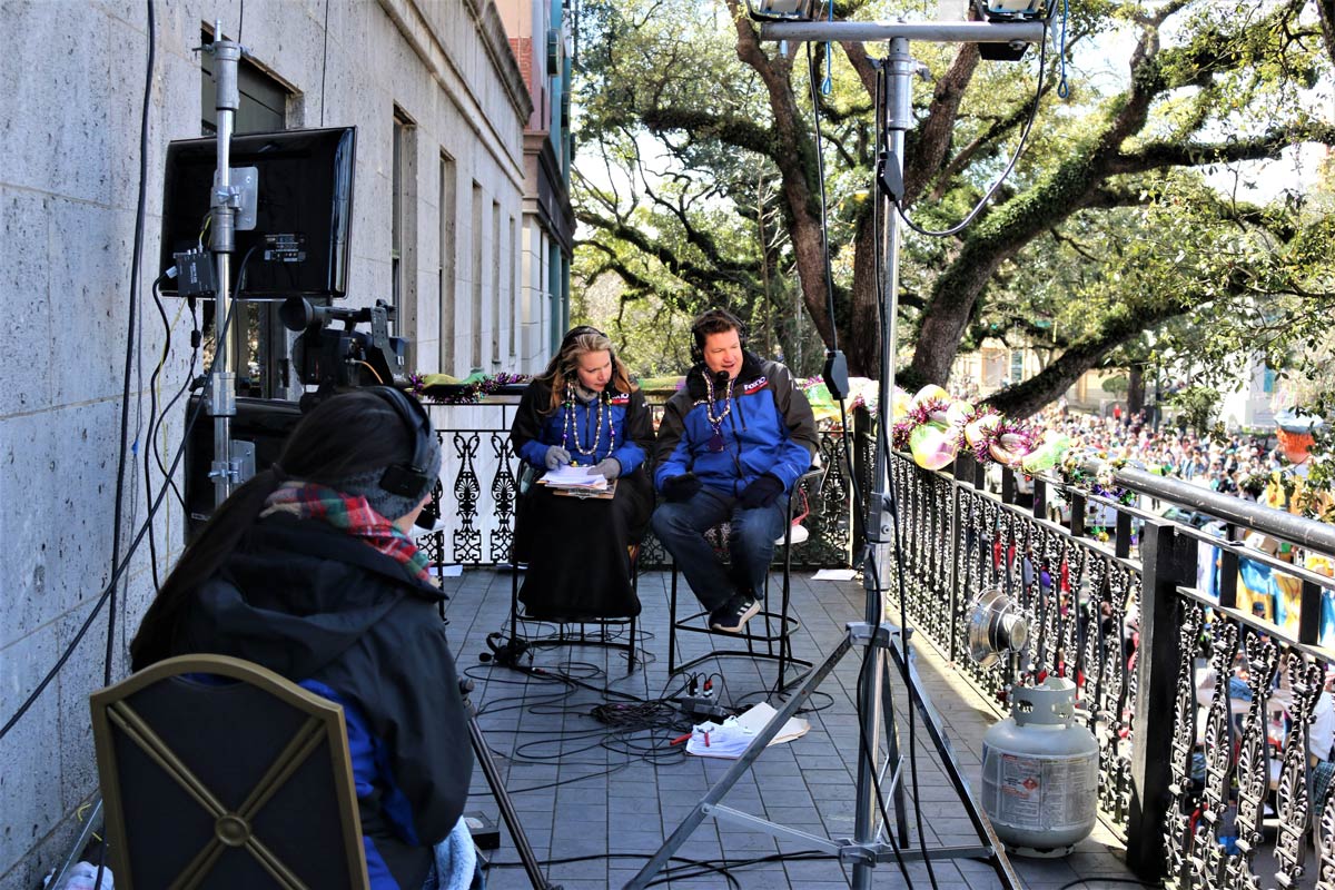 Two people on a balcony wearing blue and black jackets and headphones being filmed on a balcony