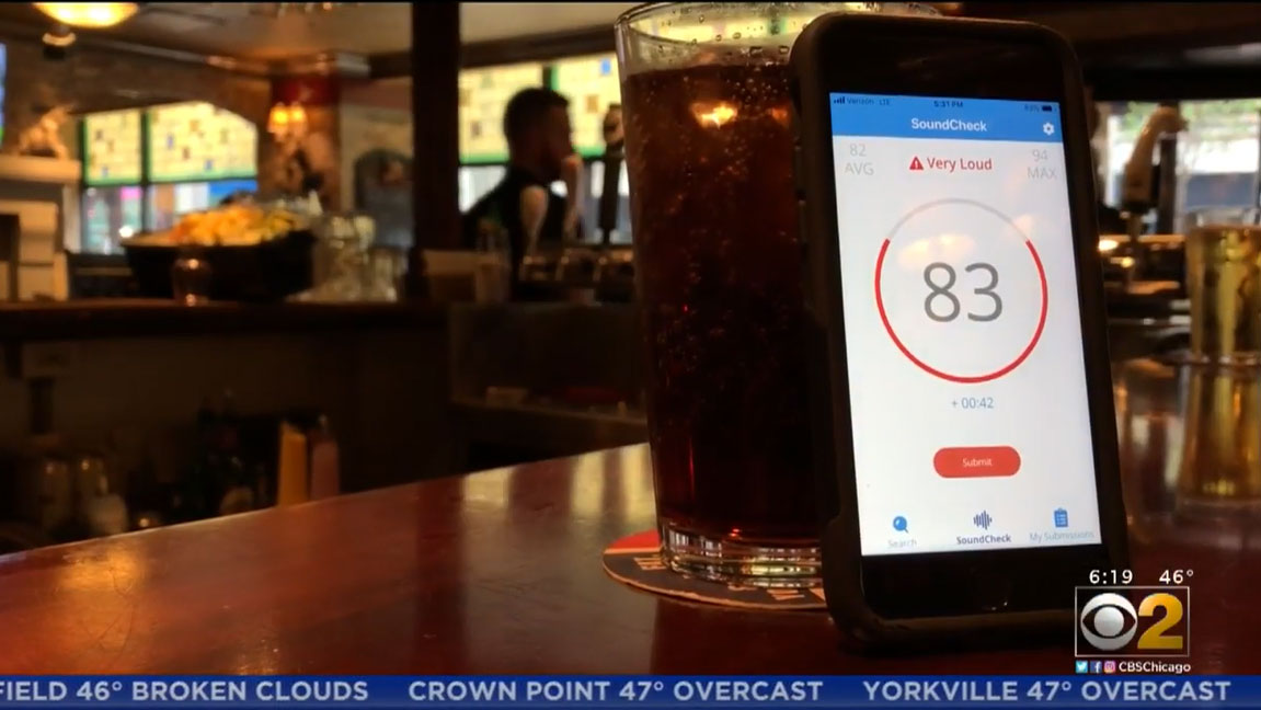 A phone showing the sound level of 83 decibels leans against a dark colored drink on a bar