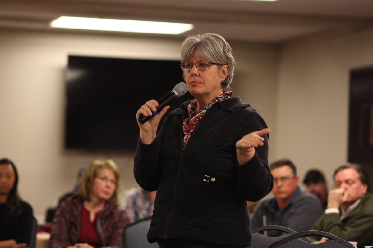 A person with short gray hair, glasses, a black jacket and a red patterned scarf stands holding a microphone during a meeting.