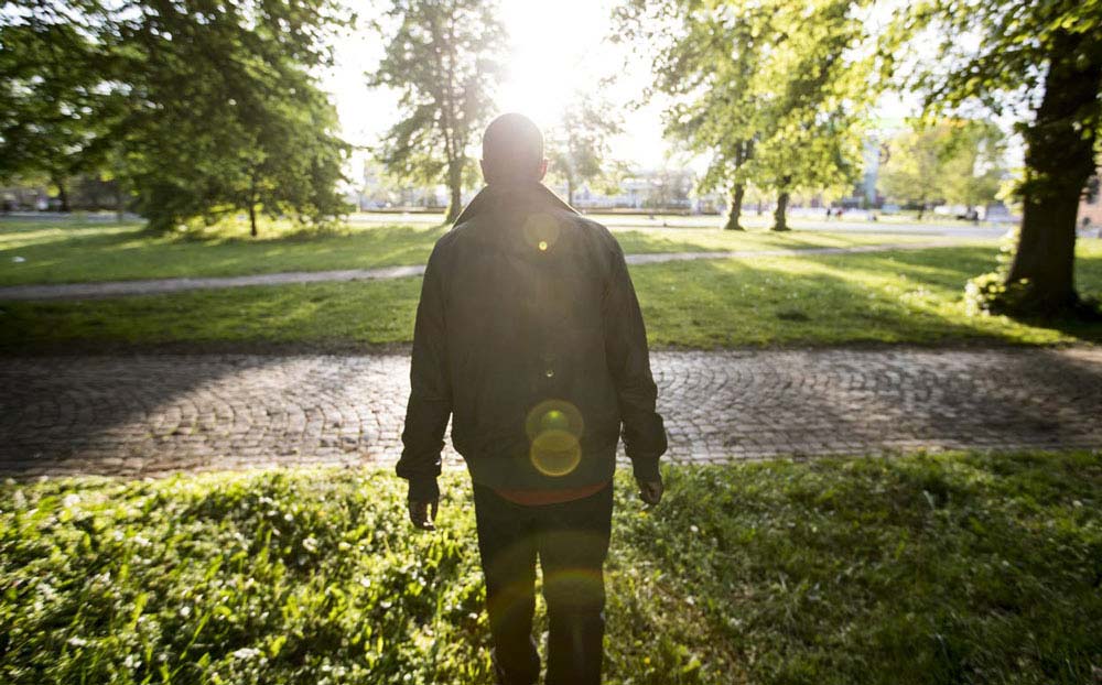 A person wearing a jacket and pants stands in a park as the sun shines on them