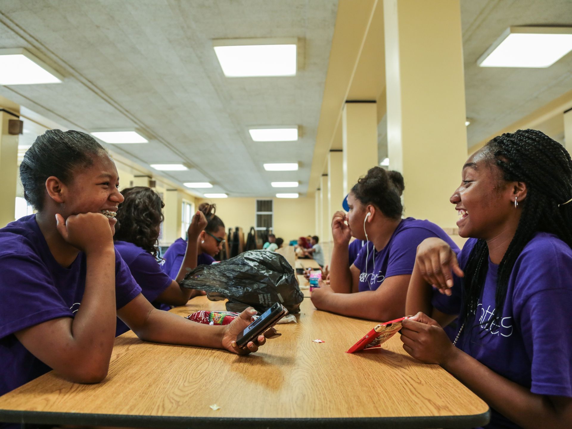 Students in blue t-shirts sitting at a table. Two in the front are laughing.