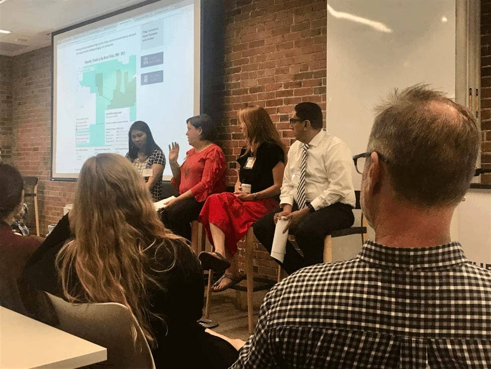 Four people seated against a brick wall while a projector plays on screen at a meeting