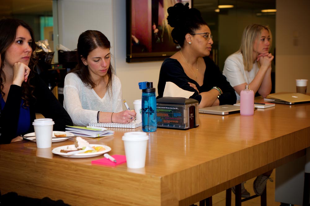 4 people seated at a wooden table. One is taking notes.