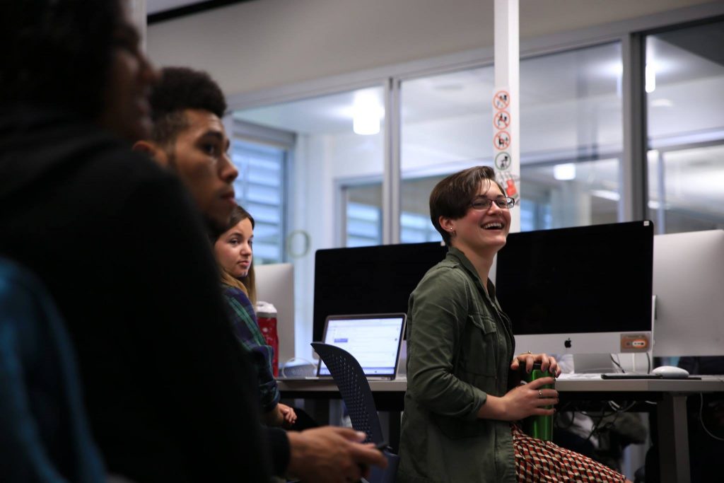 A person wearing a green button up shirt and red patterned pants sitting at a computer and smiling