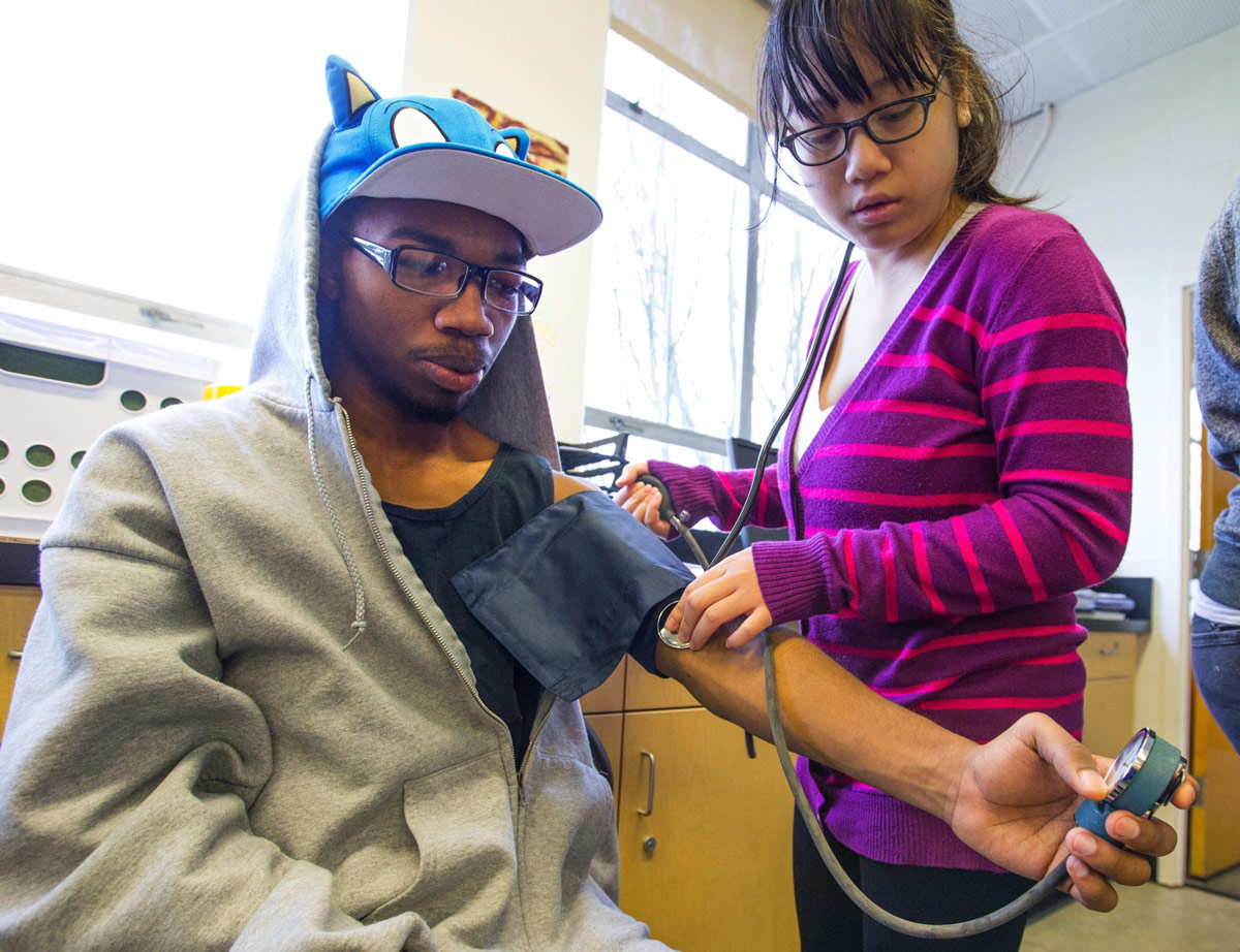 A person with glasses wearing a purple striped shirt takes the pulse of a Black student wearing glasses and a Sonic the Hedgehog hat