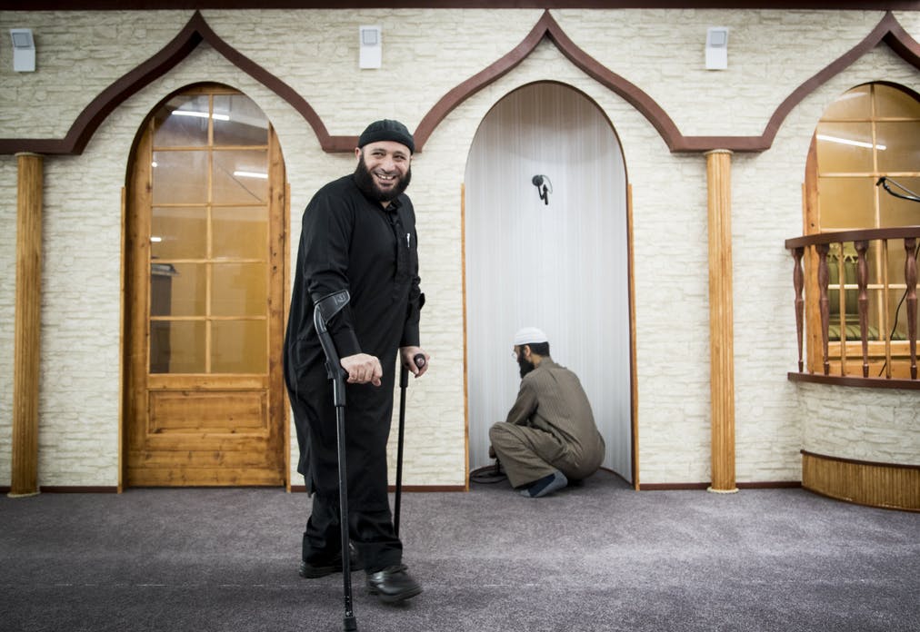 A Muslim person dressed in all black with forearm crutches smiling in a mosque.