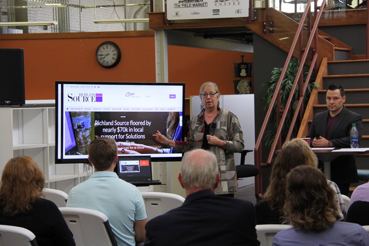 A person wearing a gray patterned shirt and glasses points to a presentation on a monitor in front of a group