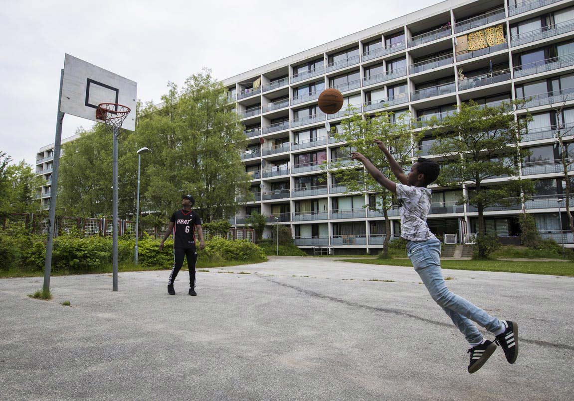 A child leaves their feet as they shoot a basketball towards the basket on a court in front of an apartment building