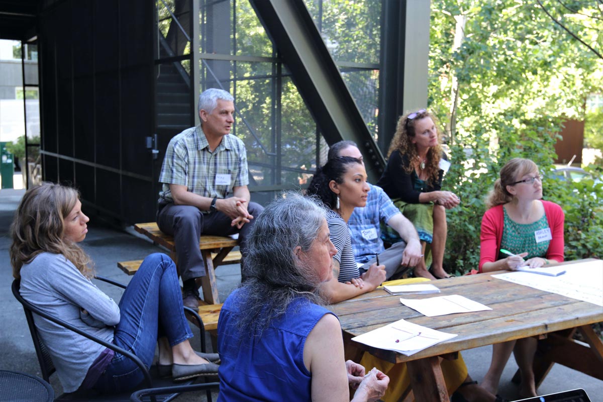 Seven people sitting at and around an outside wooden table, all looking in the same direction