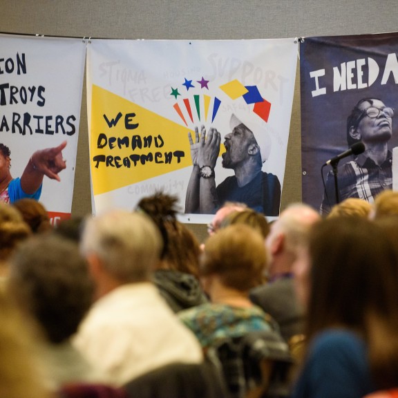 Three posters have the text "Education Destroys Barriers", "We Demand Treatment", and "I Need A Chance"