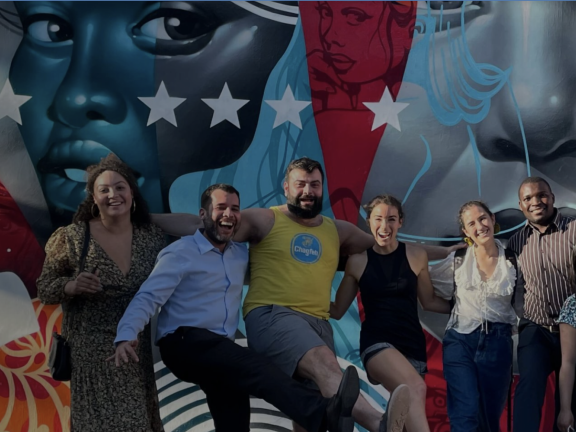 People smile and pose in front of a colorful mural