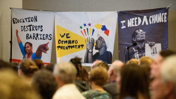 Three posters have the text "Education Destroys Barriers", "We Demand Treatment", and "I Need A Chance"